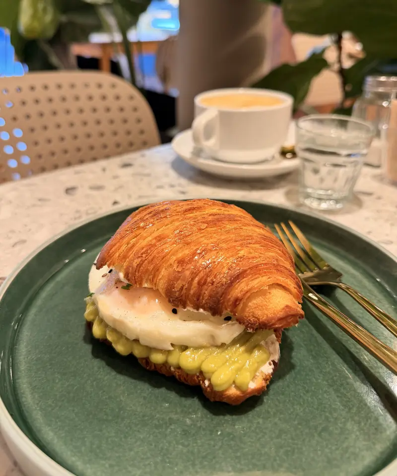 Croissant relleno de queso crema, palta y huevo, con un café con leche atrás